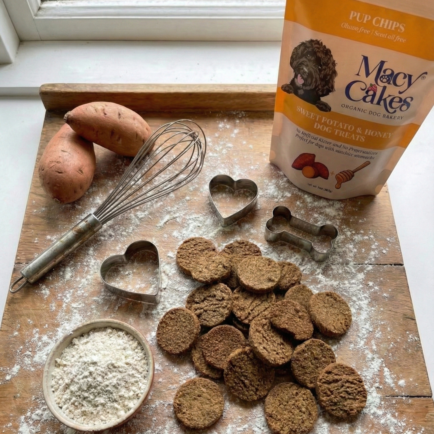 Wooden cutting board with dog treats, flour, cookie cutters, and a bag of dog food.