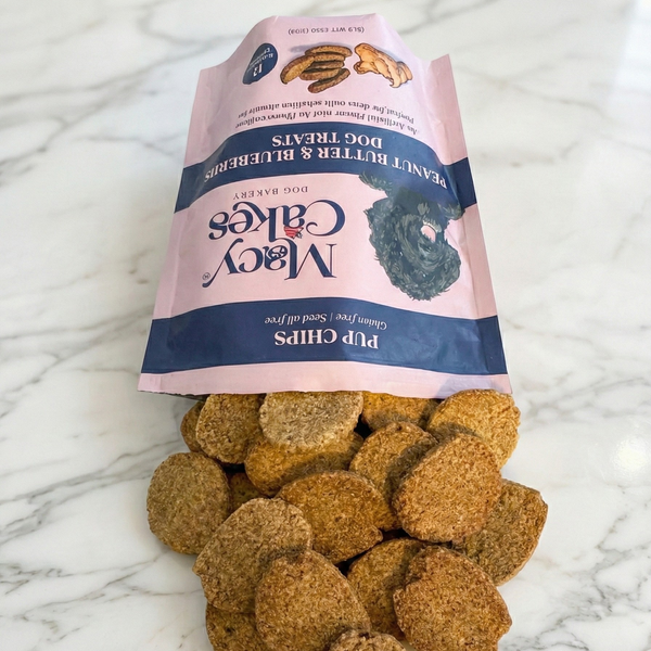 A pink and navy blue Macy Cakes Dog Bakery Pup Chips Peanut Butter & Blueberries Dog Treats bag featuring a black poodle illustration, spilling golden-brown textured oval treats onto a white marble countertop