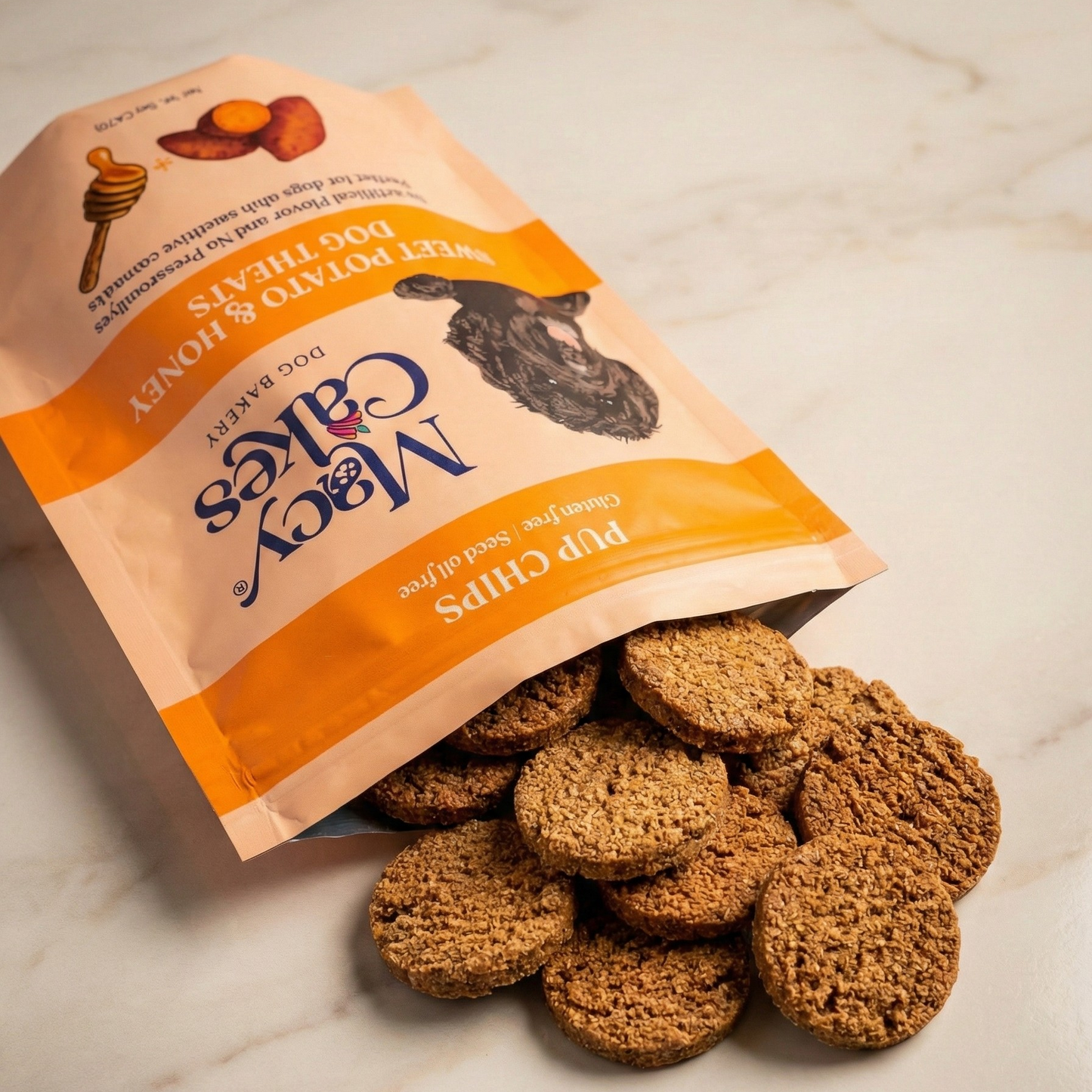 Product photography of Macy Cakes Dog Bakery Sweet Potato & Honey Pup Chips spilling from an open peach-colored bag featuring illustrations of a honey dipper, sweet potato, and a black dog's head, onto a light marble surface revealing a pile of round, textured brown treats