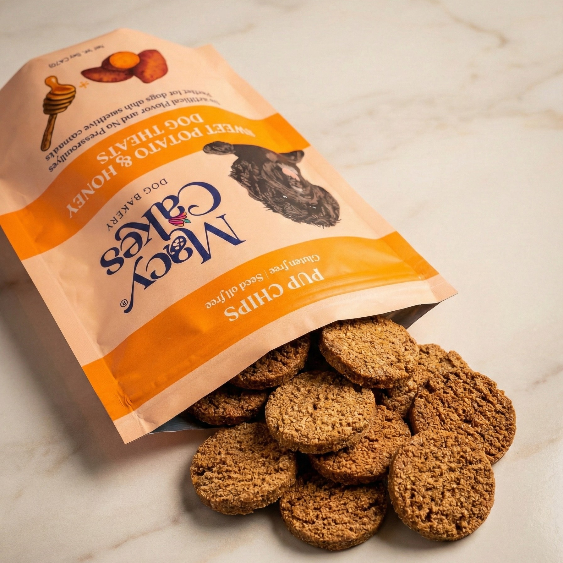 Product photography of Macy Cakes Dog Bakery Sweet Potato & Honey Pup Chips spilling from an open peach-colored bag featuring illustrations of a honey dipper, sweet potato, and a black dog's head, onto a light marble surface revealing a pile of round, textured brown treats
